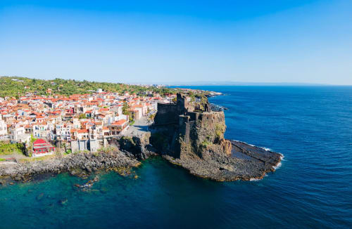 Aci Castello, villaggio costiero con edifici dai tetti rossi e una drammatica fortezza vulcanica rocciosa che domina il turchese