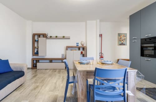 Moderna zona pranzo dell'appartamento di Brindisi con sedie blu, tavolo in legno, cucina integrata e zona living con elementi di arredo