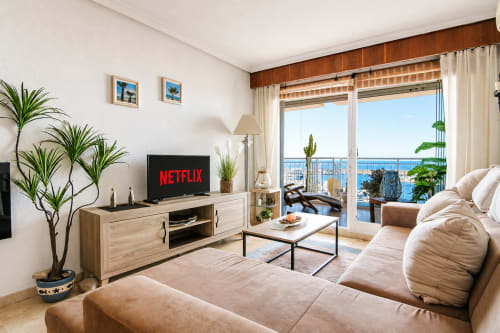 Sala de estar de la Marina Suite en Riurau Home Calpe con balcón con vista al mar, sofá beige, TV, plantas y mobiliario moderno