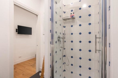 Modern bathroom at Suite Maradona Naples with blue-accented white tile shower, glass enclosure, and TV mounted on wall