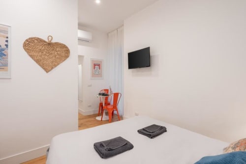Modern bedroom at Suite Maradona Naples with white walls, double bed, flat-screen TV, woven heart decoration, and orange