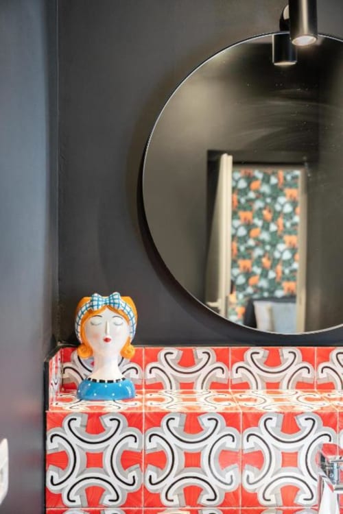Modern bathroom vanity with decorative red patterned tiles, round black mirror, and whimsical figurine at Suite Maradona