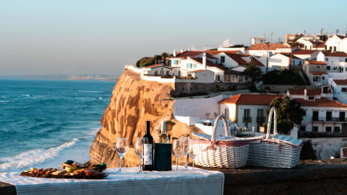 Coastal picnic setup with wine and glasses overlooking Sintra's clifftop village and turquoise ocean