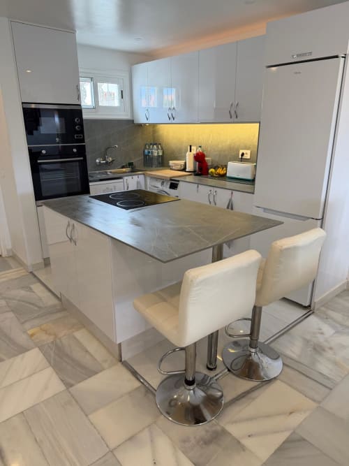 Modern white kitchen with gray countertops, stainless steel appliances, and cream bar stools at island in Costa Adeje