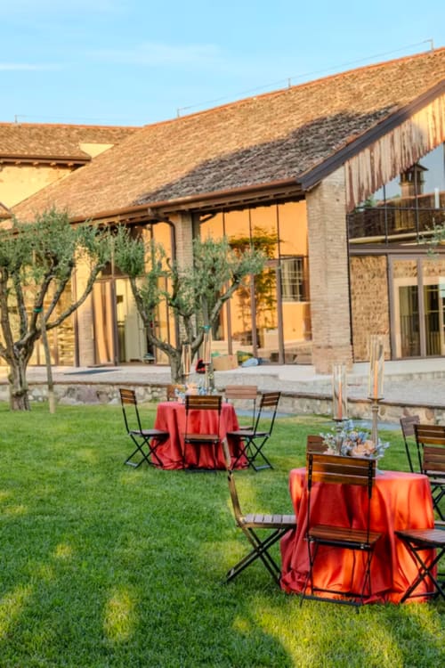 Allestimento per cena all'aperto a Corte Baruzzo Relais con tavoli rossi, ulivo e rustica costruzione in pietra a Volta