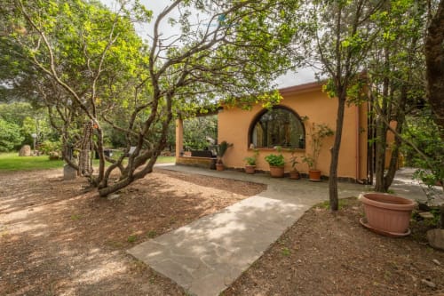 Giardino di Villa Chia con alberi maestosi, percorso in pietra, vasi in terracotta e ingresso della villa dipinto d'arancio a Quartu
