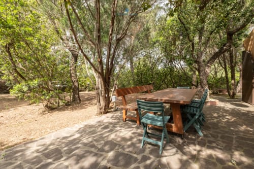 Zona pranzo all'aperto con tavolo in legno e sedie verdi sotto gli alberi ombreggiati di Villa Chia, Quartu Sant'Elena