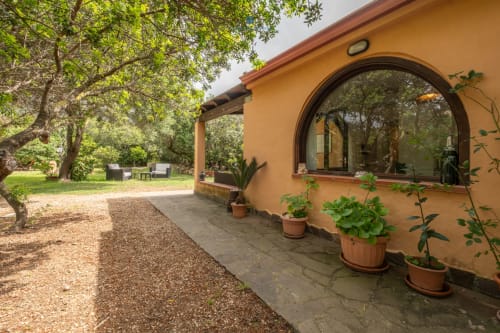 Villa Chia con muri color terracotta, grande finestra ad arco, piante in vaso, vialetto di ghiaia e lussureggiante giardino