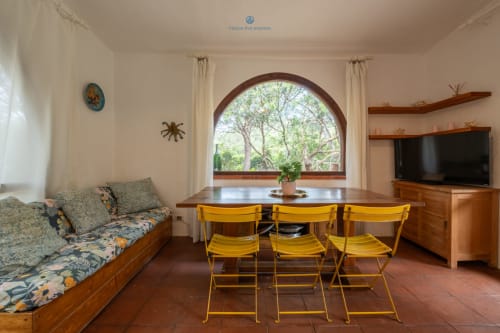 Soggiorno di Villa Chia con sedie gialle, tavolo in legno, divano, TV e finestra ad arco con vista sul giardino a Quartu Sant'elena