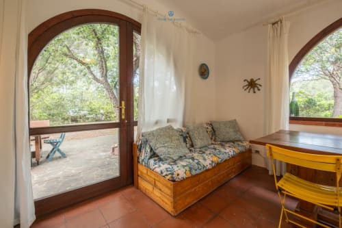 Camera da letto luminosa con letto in legno, sedia gialla e finestre ad arco con vista sul giardino - House Live Express, Quartu