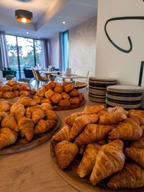 Croissants frais exposés sur des planches en bois dans la salle petit-déjeuner de l'hôtel La Verriaire avec coin repas moderne et grandes fenêtres