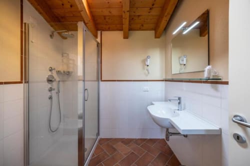 Bagno moderno presso il Corte Baruzzo Relais con doccia aperta, lavabo bianco, ampio specchio, soffitto in legno e pavimento in cotto