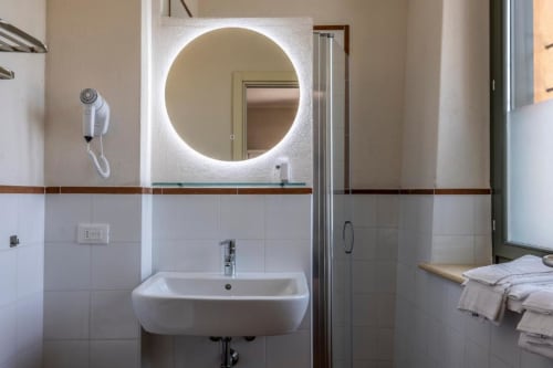 Bagno moderno della Corte Baruzzo Relais con specchio rotondo illuminato, lavabo bianco e doccia con divisorio in vetro nella camera Comfort