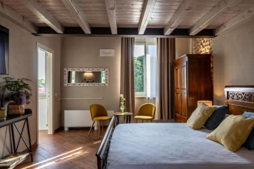 Elegante suite presso Corte Baruzzo Relais con travi in legno, letto matrimoniale, zona soggiorno e vista sul giardino attraverso pavimento in cotto