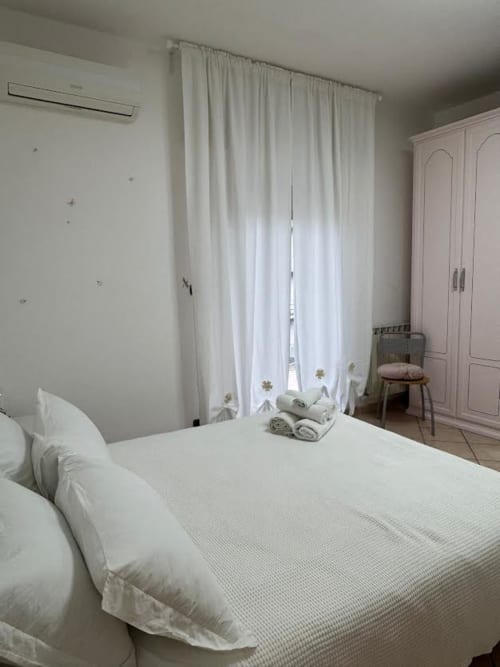 Camera da letto luminosa dell'appartamento a Catania con letto bianco, tende trasparenti, armadio rosa e arredi moderni