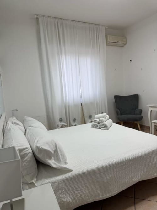 Camera da letto moderna e luminosa presso Catan Appartament con letto bianco, asciugamani piegati, tende trasparenti, poltrona scura e aria condizionata