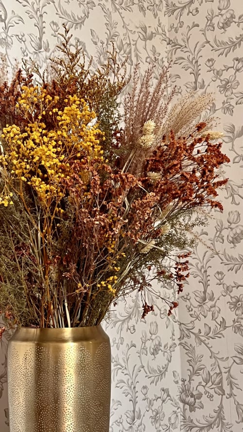 Composition de fleurs séchées dans un vase doré devant un papier peint à motifs floraux au Domaine du Faure
