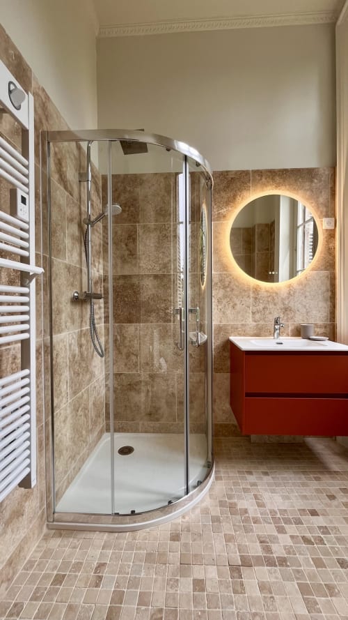 Salle de bains moderne avec douche en verre incurvé, carrelage beige, vanité rouge et miroir rond illuminé au Domaine du