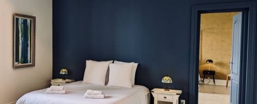 Chambre élégante au Domaine du Faure aux murs bleu marine, literie blanche, lampes jumelées et vue sur le jardin par la porte