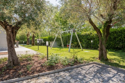 Giardino di Corte Garda con alberi maturi, patio pavimentato, altalena e prato curato a Serniga