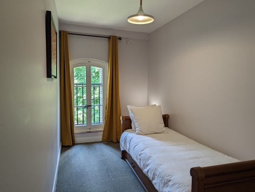 Lit simple dans une chambre lumineuse du Domaine du Faure avec vue sur le jardin par des portes cintrées et rideaux moutarde