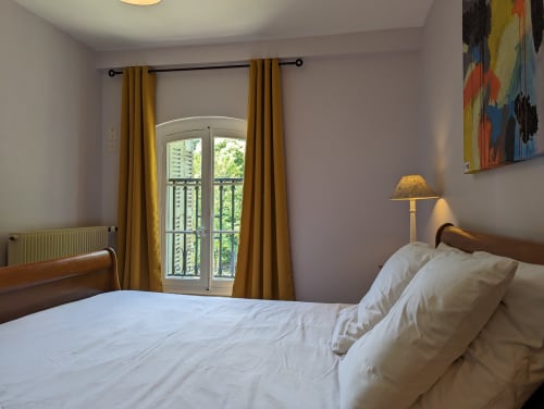 Chambre lumineuse avec literie blanche, lit en bois, rideaux moutarde, portes-fenêtres sur jardin et œuvres d'art colorées au Domaine du