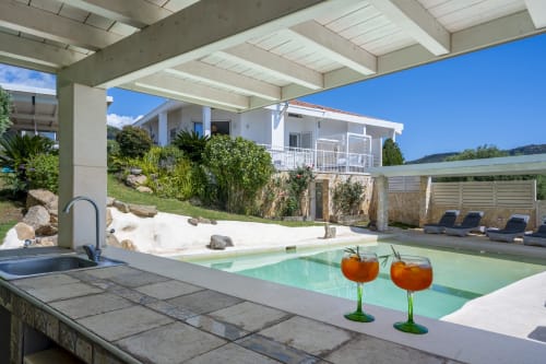 Luxury villa pool at Top Villas Sardinia with cocktails, concrete beams, white house, and mountain views