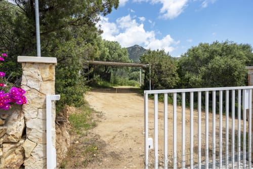 White gate entrance to Top Villas Sardinia with stone pillar, magenta bougainvillea, dirt driveway, and mountain views in