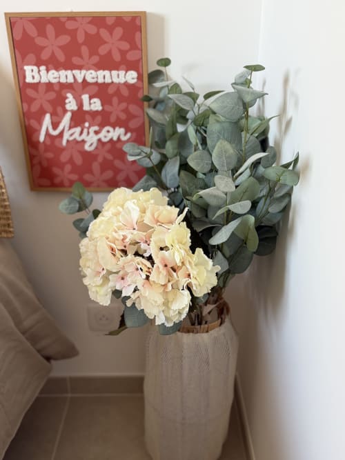 Bouquet d'hortensias crème avec eucalyptus dans un vase blanc, à côté d'une pancarte rouge 'Bienvenue à la Maison' au Petit Coin de