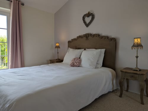 Chambre élégante avec tête de lit ornée, literie blanche, accents rose, décor cœurs, vue sur jardin par rideaux rose