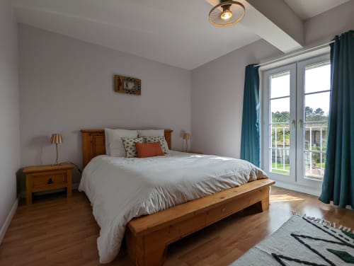Chambre lumineuse avec lit en bois, portes-fenêtres donnant sur le jardin, rideaux bleu-canard et équipements modernes au gîte Domaine du Faure