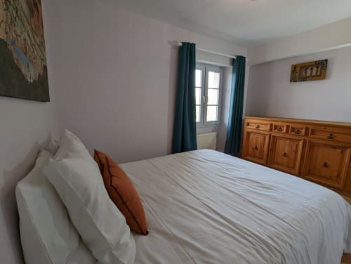 Chambre lumineuse avec lit queen, draps blancs, commode en bois, rideaux bleu-vert et vue sur le jardin du gîte Domaine du Faure