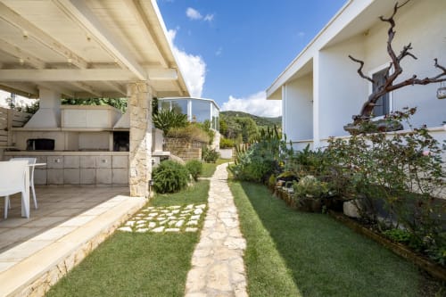 Modern villa in Sardinia with stone pathway, manicured gardens, outdoor kitchen, and sea views from private patio area