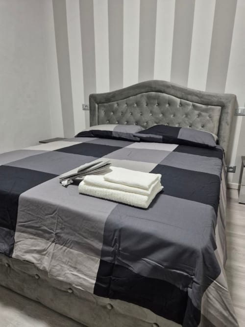 Camera da letto moderna a Mia Monza Living con testata imbottita grigia, carta da parati a strisce e biancheria letto color block con asciugamani