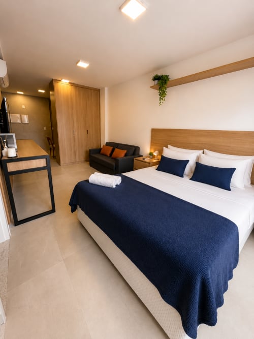 Quarto moderno com cama queen, roupa de cama azul-marinho, cabeceira de madeira, área de estar com sofá preto, escrivaninha e teto com vigas de madeira