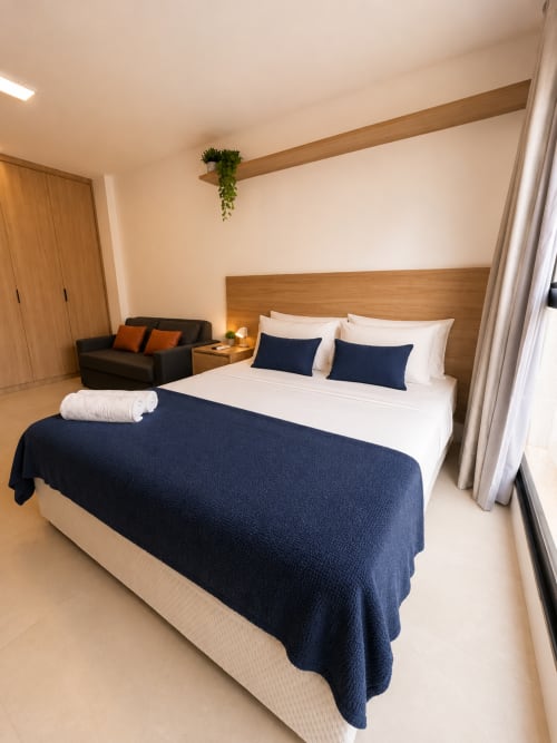 Quarto de apartamento moderno em Brasília com cama king, roupa de cama azul-marinho, cabeceira de madeira, área de estar e vistas para pontos turísticos