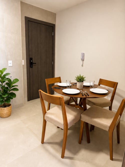 Área de refeições moderna com mesa e cadeiras de madeira, paredes bege, porta escura e planta verde em apartamento em Brasília.