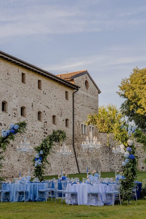 Allestimento ricevimento matrimoniale esterna presso Corte Baruzzo Relais con decorazioni floreali blu e tovaglie bianche su sfondo storico