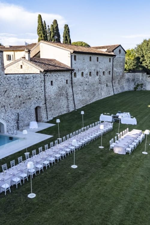 Allestimento di ricevimento di nozze all'aperto presso Corte Baruzzo Relais con sedie e tavoli bianchi e sfondo di fortezza in pietra a Volta