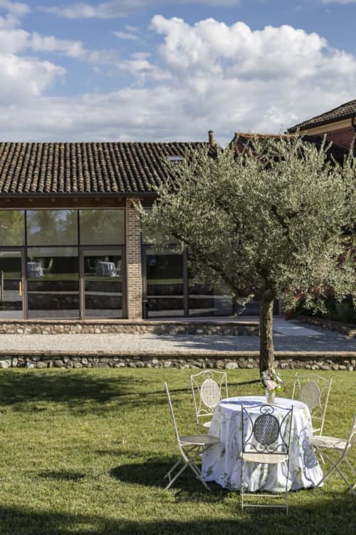 Zona pranzo esterna al Corte Baruzzo Relais con tavolo elegante, sedie, ulivo e moderno edificio in laterizio a Volta