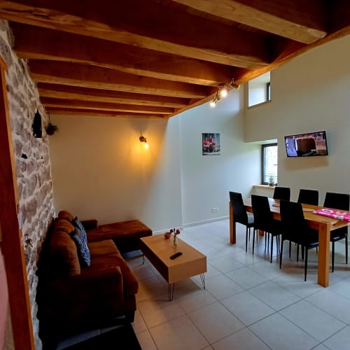 Séjour et salle à manger ouverts aux Gîtes Du Couvent avec poutres apparentes, canapé marron, table en bois et télévision