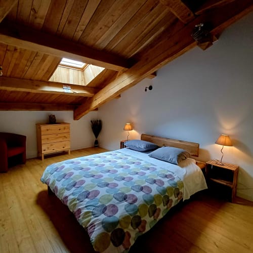 Chambre spacieuse aux poutres apparentes, lit queen-size, tables de nuit en bois et lumière chaleureuse aux Gîtes Du Couvent