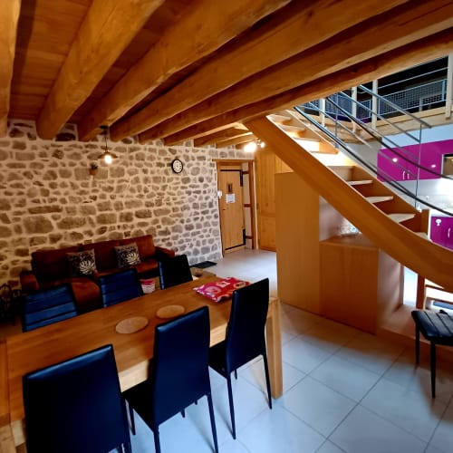 Espace ouvert de salle à manger et séjour avec poutres en bois, murs en pierre, chaises bleues, table en bois et escalier aux Gîtes Du