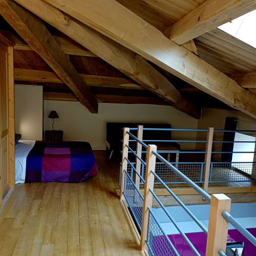 Chambre en mezzanine aux poutres apparentes, lit avec couverture mauve, rampes métalliques et parquet aux Gîtes Du Couvent