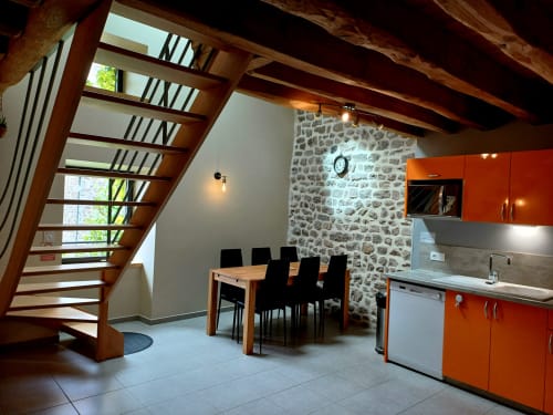 Cuisine rustique avec poutres en bois, mur en pierre, placards orange et escalier en bois aux Gîtes Du