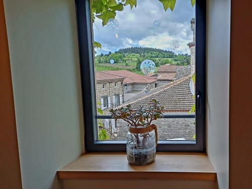 Plante en pot sur le rebord de fenêtre avec vue sur la campagne ardéchoise, bâtiments en pierre et collines ondulantes aux Gîtes Du Couvent