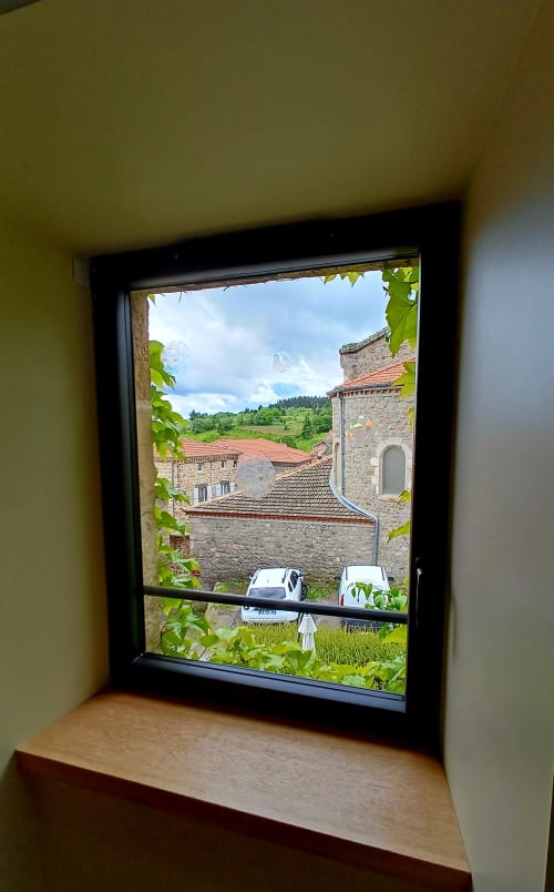 Vue depuis une fenêtre encadrée de lierre : bâtiments en pierre du village et collines verdoyantes aux Gîtes Du Couvent, refuge en Ardèche