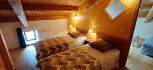 Chambre cosy des Gîtes Du Couvent avec lits jumeaux, poutres en bois, éclairage chaleureux et murs en pierre apparente en Ardèche