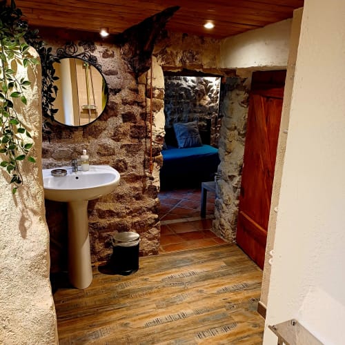 Salle de bain rustique en Ardèche dans un gîte avec murs en pierre, lavabo sur colonne, miroir rond et lit bleu visible par la porte