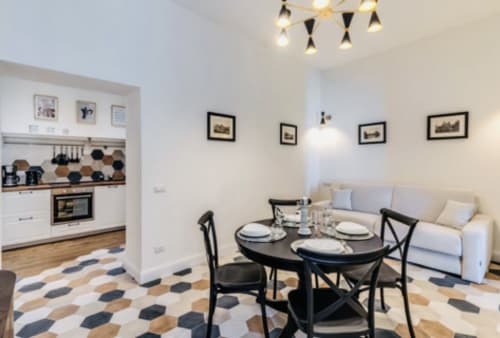 Zona pranzo di appartamento romano moderno con tavolo nero, divano color crema, cucina bianca, arte incorniciata e motivi geometrici sul pavimento